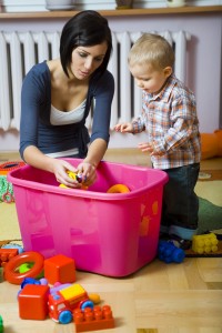 Young woman with baby boy during plaing. Woman showing toy to baby. They are at container with toys. Front view.